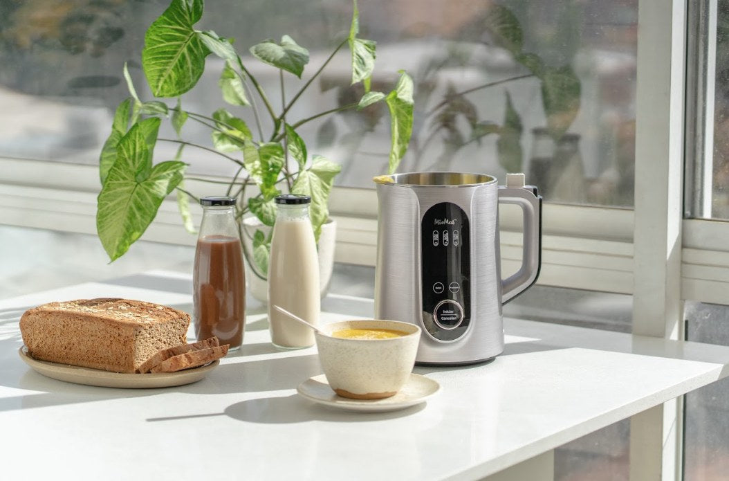 Máquina para hacer sopas MioMat Pro sobre la mesa junto a una taza de crema, pan y leches vegetales